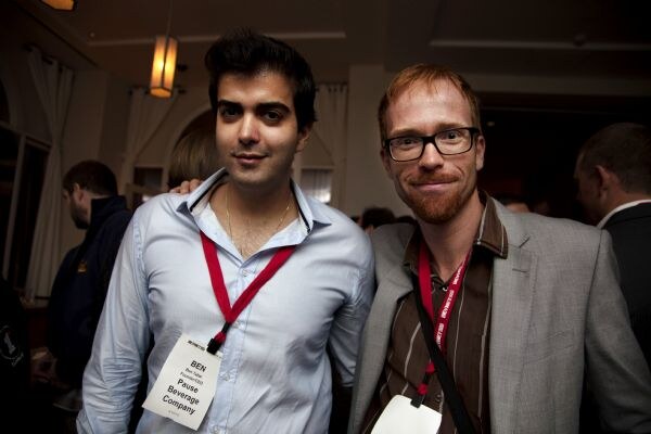 Ben of Pause Beverage Company, on the left, in a light blue shirt and red lanyard, poses beside a man with glasses and a gray blazer in a dimly lit room.