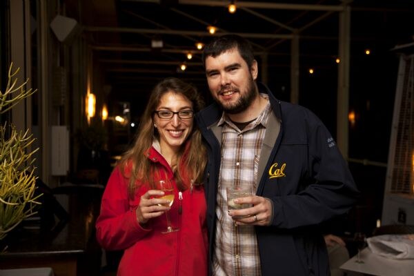 A smiling woman with glasses in a red jacket holds a glass of white wine beside a bearded man in a navy jacket with a Cal logo, holding a drink, with string lights behind them.
