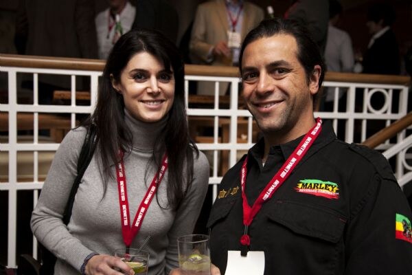 A smiling woman in a gray turtleneck stands beside a man in a black shirt with a Marley logo. Both wear red lanyards and hold drinks in front of a white railing.