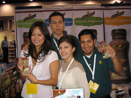 Four people in a group pose closely at a booth with Taste Nirvana banners behind them. Two of them hold small bottles of the product.