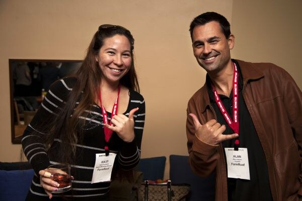 Kalei and Alan of FarmRoof smile and make shaka hand gestures. Both wear red lanyards with name badges. Kalei holds a drink, and Alan wears a brown leather jacket.