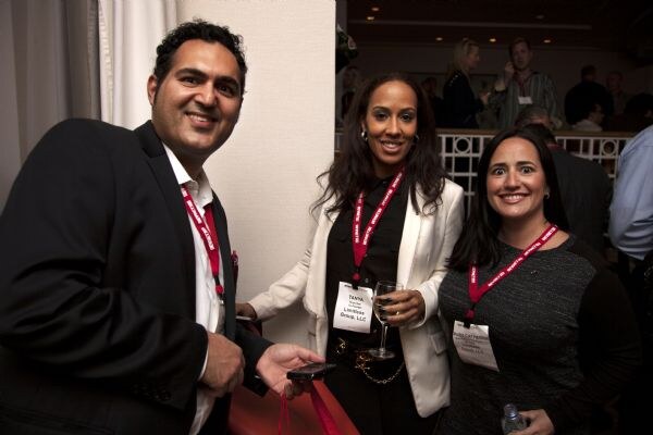 Tanya, in the center, wears a white blazer and holds a wine glass. A smiling man in a dark suit stands at left and a woman in a gray top at right. All wear red lanyards.