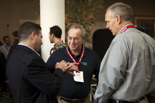 Sandy, center, wears a navy polo and red lanyard and listens to a gray-haired man in a gray shirt at right, while a man in a dark jacket at left gestures with his hands.