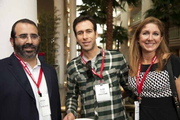 Three attendees with red lanyards pose together indoors. Serge, a bearded man in glasses and dark blazer, stands left; Elan of Coco Cafe, in a green plaid shirt, is center; a woman stands right.