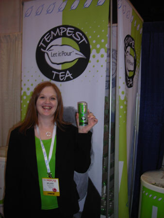 A smiling woman with long reddish hair, a black jacket and a green top holds up a can of Tempest Tea in front of green and white Tempest Tea banners.