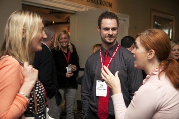 Rob of Virginia Dare, wearing a gray shirt, red tie and red lanyard, stands in the center smiling as two women in the foreground talk. Other attendees stand behind near a ballroom entrance.