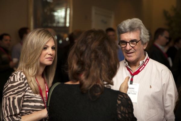 Harry, a gray-haired man in glasses and a striped shirt with a red lanyard, stands at right talking with a blonde woman in a patterned top and a woman seen from behind.