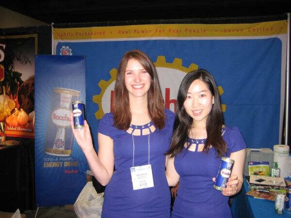 Two smiling women in matching blue tops each hold a can of Bacchus energy drink in front of a Bacchus banner and a tall Bacchus can display in the booth.