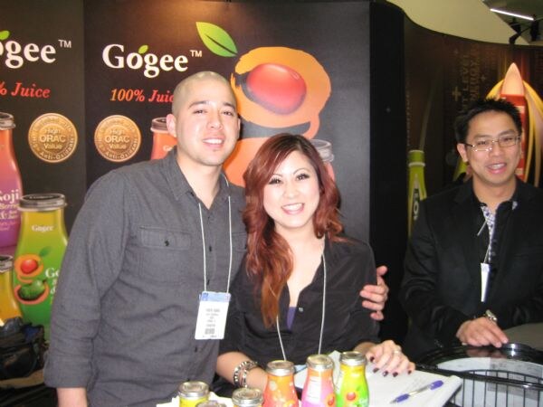 Two men and a woman stand behind a counter in front of Gogee 100% juice banners. Bottles of juice sit on the counter in front of them, and two of them wear badges on lanyards.