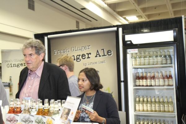 A gray-haired man in a dark jacket and a woman in a navy blazer stand at the Bruce Cost Ginger Ale booth, with bottles on the counter and a lit refrigerator of ginger ale behind them.