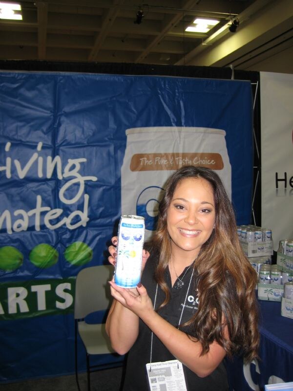 A smiling woman with long brown hair, wearing a black C2O shirt and a badge, holds up a can of C2O coconut water in front of a blue banner. Cans are stacked on a table behind her.