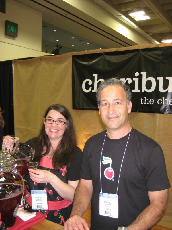 A woman with glasses holding a sample cup and a man in a black shirt with a cherry logo stand at the Cheribundi booth, beside a glass pitcher of red juice and a partly visible Cheribundi banner.