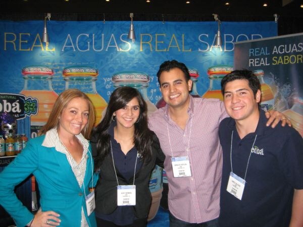Four smiling people stand close together in front of a blue Cobá banner reading "Real Aguas Real Sabor", with large juice bottle graphics behind them. Three wear badges on lanyards.