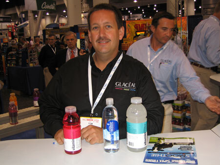 Tom, wearing a black Glacéau shirt and lanyard, sits at a table with vitaminwater and smartwater bottles and a Beverage Spectrum magazine. Two men stand behind him.
