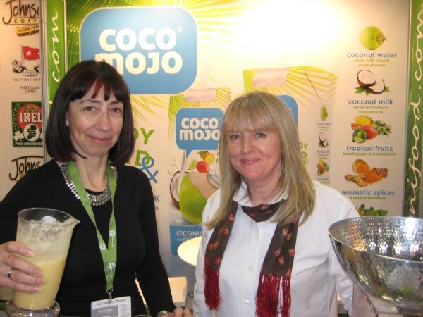 Two women stand smiling in front of a Coco Mojo display; one holds a pitcher of pale drink, and a banner lists coconut water, coconut milk, tropical fruits and aromatic spices.
