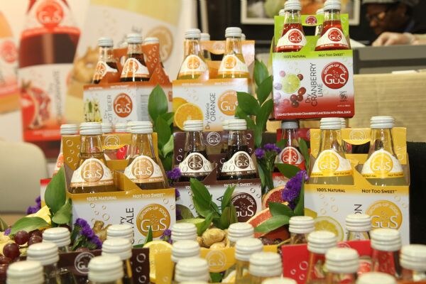 Stacked cardboard carriers of GuS soda bottles, including Dry Orange and Cranberry Lime, are arranged with green leaves, purple flowers and fruit.