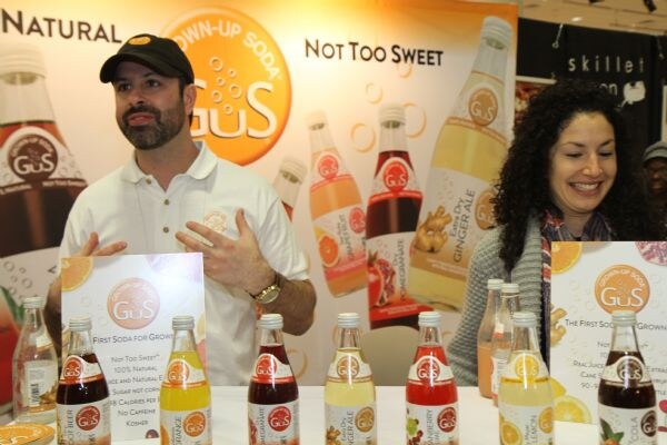A bearded man in a cap and a smiling woman with dark curly hair stand behind a table of GuS soda bottles and signs reading Not Too Sweet, with a large GuS banner behind them.