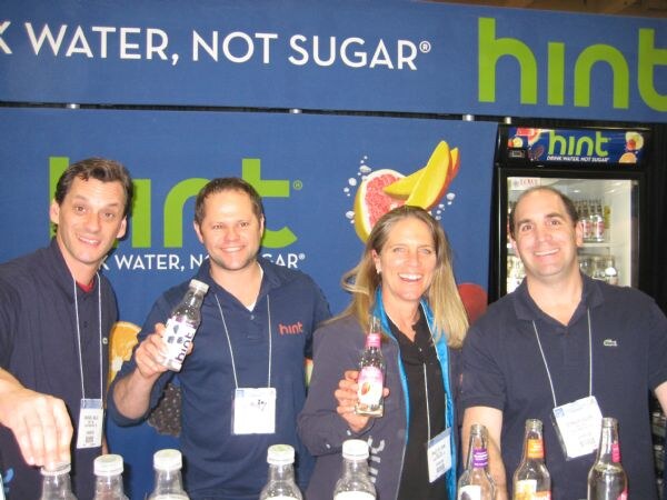 Four people in front of a Hint banner reading Drink Water, Not Sugar pose behind a counter; two hold bottles of flavored Hint water.