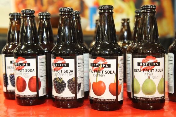 A row of brown glass Hotlips Real Fruit Soda bottles on a red surface, with labels for cranberry, black raspberry, cherry and pear.
