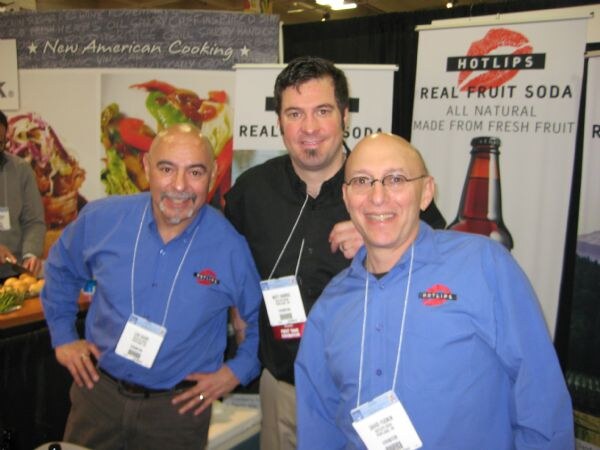 Three smiling men wearing badges stand together in front of Hotlips Real Fruit Soda banners; two wear blue shirts with the Hotlips logo and one wears black.