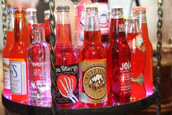A lit display of glass soda bottles in red, orange and clear liquids, including Kutztown Ginger Beer and Joia sodas, plus a Love Potion No. 9 bottle.