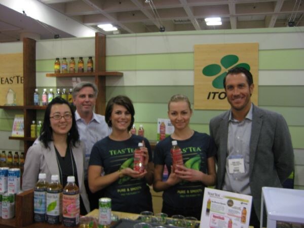 Five people stand behind a counter at a Teas' Tea booth with an Ito En sign; two women in Teas' Tea shirts hold bottles of tea, and bottles and cans are on display.