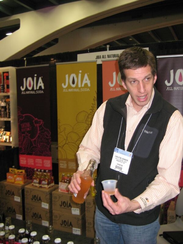 Carleton Johnson, wearing a Marmot vest and exhibitor badge, holds a bottle of soda and a sample cup in front of Joia All Natural Soda banners and stacked cases.
