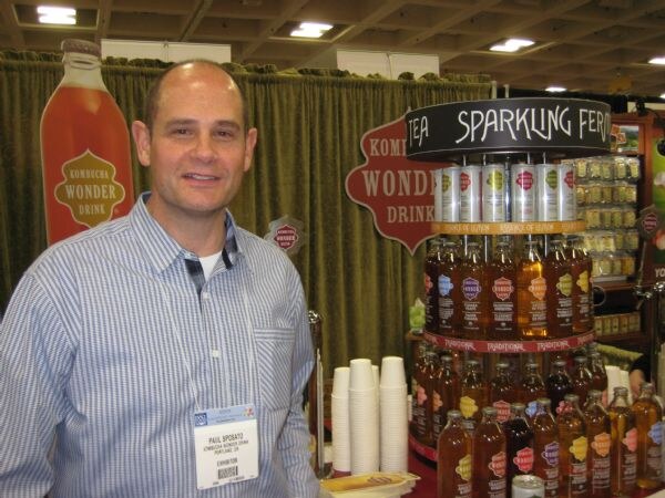 Paul Sposato of Kombucha Wonder Drink stands smiling in a striped shirt beside a display rack of Kombucha Wonder Drink bottles and cans.