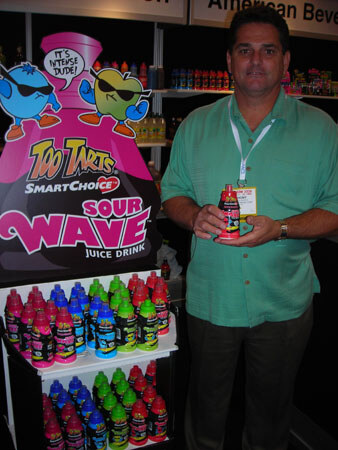 A man in a green shirt holds a red bottle beside a Too Tarts SmartChoice Sour Wave juice drink display with rows of colorful bottles and cartoon characters.