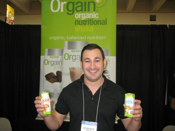 Matthew Pauling of Orgain smiles at the camera in a black polo, holding a small Orgain Kids carton in each hand in front of an Orgain organic nutritional shake banner.