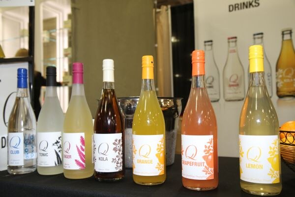 Eight Q Drinks bottles stand in a row on a black-draped table, labeled Club, Tonic, Ginger, Kola, Orange, Grapefruit and Lemon, with more bottles pictured on a banner behind.