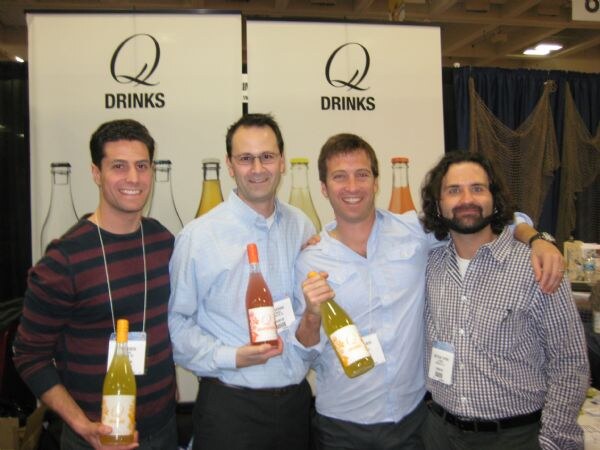 Four smiling men stand together in front of Q Drinks banners, two of them holding bottles of Q Drinks, with the arms of some around the shoulders of others.