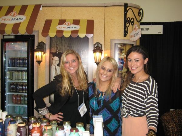 Three smiling young women stand together in front of a Real Beanz booth with striped awnings, a refrigerated display case on the left and jars of product on the counter in front.