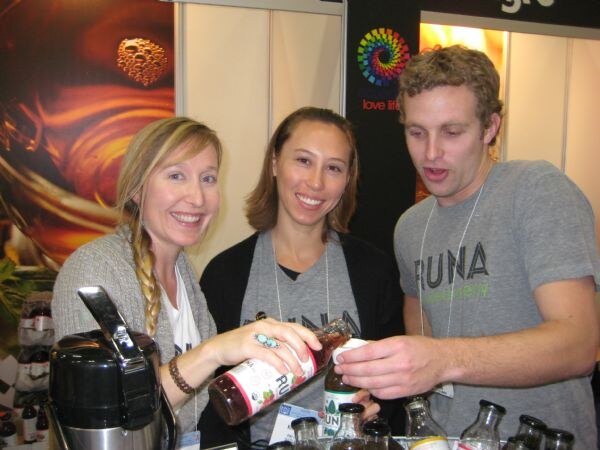 Two women and a man at a Runa booth look at a bottle of Runa drink held by the woman on the left, with a carafe in the foreground. The two on the right wear gray Runa shirts.