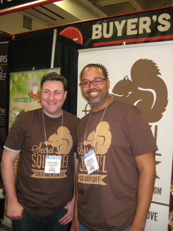 Patrick O'Donnell, left, and Trevor Smith, right, stand smiling in brown Secret Squirrel Cold Brew Coffee T-shirts in front of a banner with a large squirrel graphic.