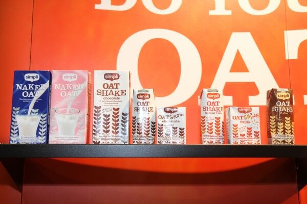 Simpli oat drink cartons, including Naked Oat, Oat Shake and Oat Grow varieties, sit in a row on a black shelf against an orange wall with large white lettering.