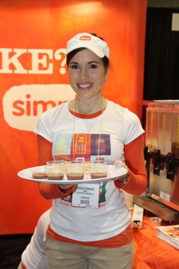 Jessica Bernard of Simpli smiles in a white Simpli visor and T-shirt, holding a tray of small cups of chocolate-colored drink samples in front of an orange Simpli banner.