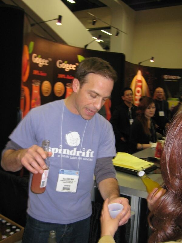 Bill Creelman of Spindrift, in a Spindrift T-shirt, holds a bottle in one hand and pours a sample into a small cup held by a person in the foreground, with Gogee signs behind him.