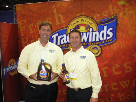 Two smiling men in yellow Tradewinds shirts and badges stand in front of a Tradewinds brewed tea backdrop; one holds a large jug and the other a bottle of tea.