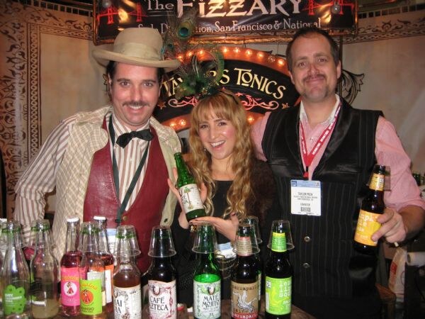 Two men and a woman smile behind a counter lined with bottled sodas under The Fizzary and Taylor's Tonics signs. The woman and one man hold bottles; one man wears a hat and bow tie.