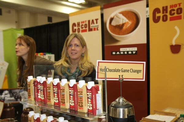 A smiling blonde woman stands behind a glass display of The City Bakery hot chocolate cartons, beside a sign reading Hot Chocolate Game Changer. Another woman looks away behind her.