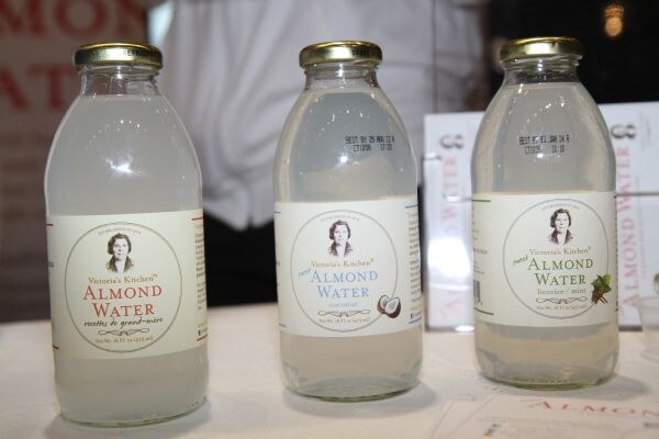 Three glass bottles of Victoria's Kitchen Almond Water on a table, labeled original, coconut and licorice mint, each with a portrait of a woman on the label.