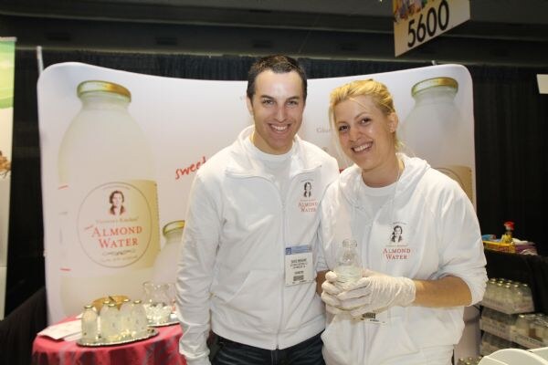 A man and a woman in white Almond Water jackets smile in front of a Victoria's Kitchen backdrop. The woman wears gloves and holds a small bottle; a table with glasses is at left.