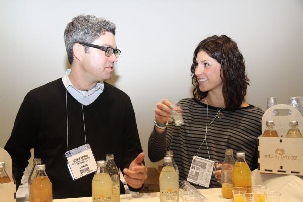 A man with glasses and a woman with dark curly hair talk at a table with bottles of golden juice and a wooden carrier marked wild poppy. The woman holds a small sample cup.
