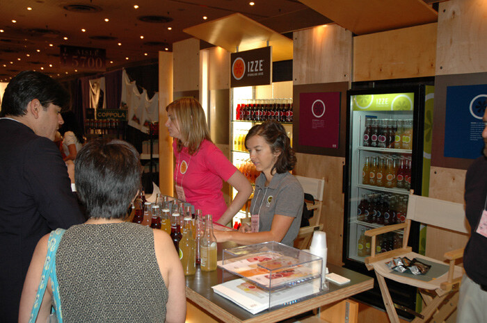 Two women in IZZE shirts staff a wooden booth counter with bottles of sparkling juice on display. Two visitors stand in the foreground, and a lit cooler of IZZE drinks is behind.