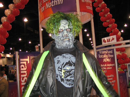 A person in a green-haired zombie mask and black jacket with neon green straps stands in front of red TransFund ATM signs and red balloon decorations on a trade show floor.