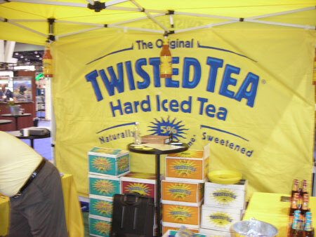 A yellow canopy booth with a large Twisted Tea Hard Iced Tea banner, stacked product cartons and a small table, with a person partly visible at the left edge.