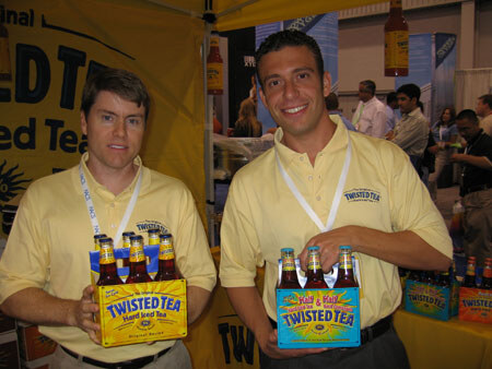 Two men in yellow Twisted Tea polo shirts each hold a six-pack of Twisted Tea bottles in front of a yellow Twisted Tea banner.