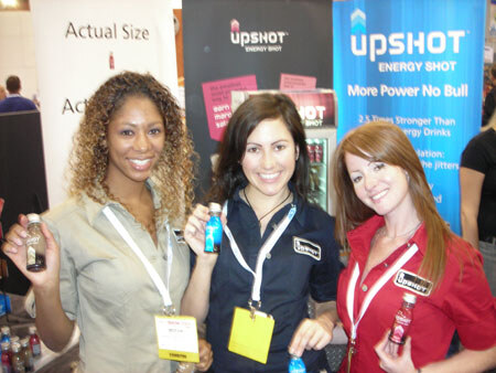 Three smiling women with lanyards hold small energy shot bottles in front of Upshot Energy Shot banners reading 'More Power No Bull'.