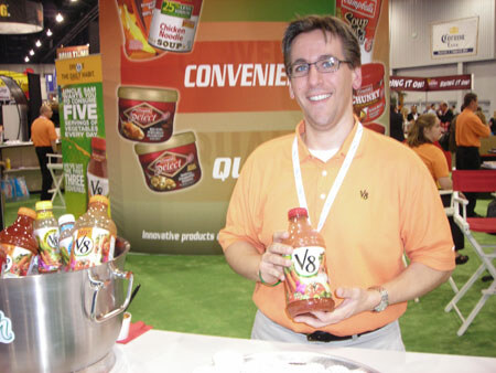 A smiling man in glasses and an orange shirt holds a bottle of V8 beside a bucket of V8 bottles, with Campbell's Select Harvest soup graphics and a Corona sign behind him.